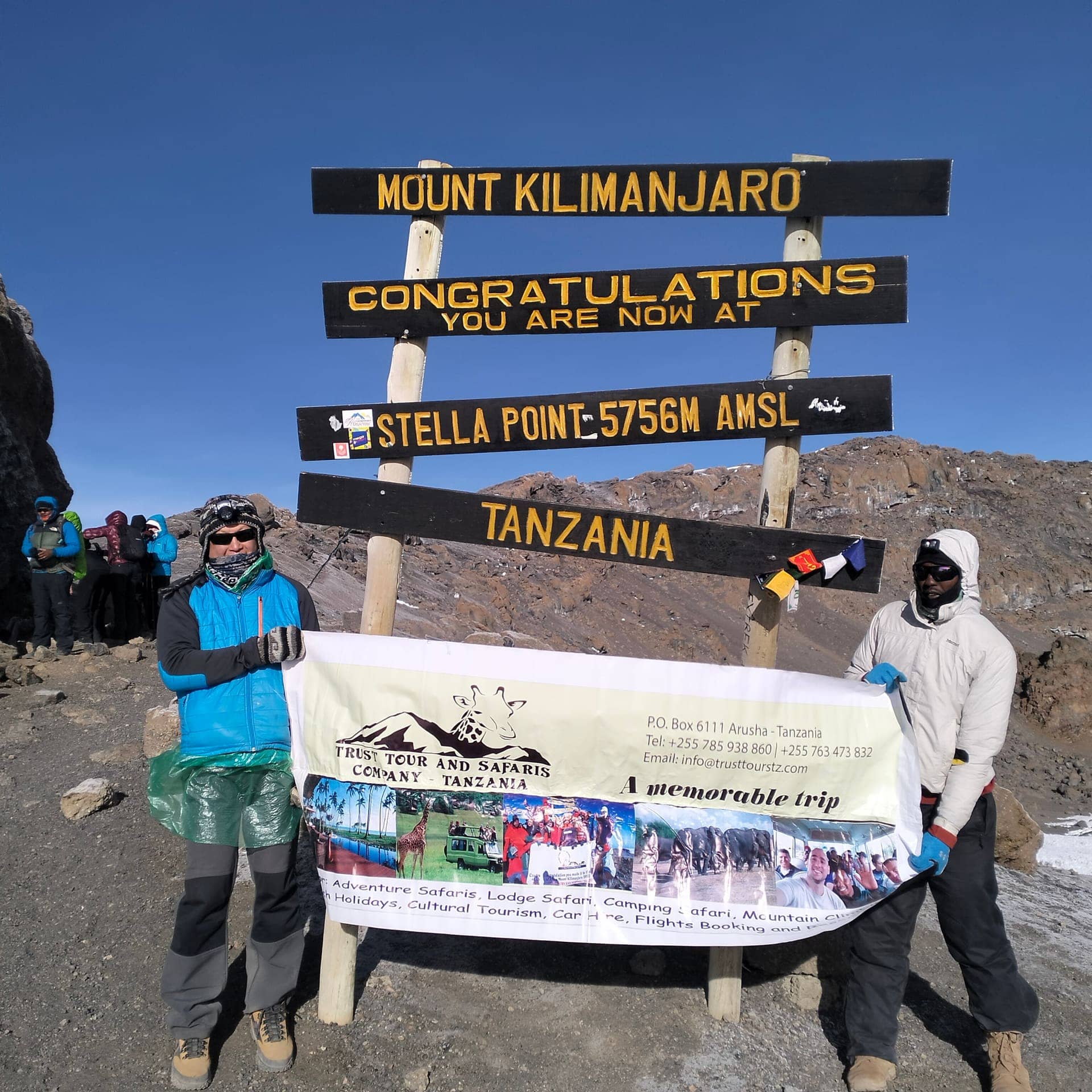 A climber and guide with the Trust Tours banner at a Kilimanjaro summit sign
